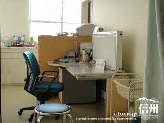 明るい部屋で医師の机と椅子、患者さんの椅子と持ち物籠が用意